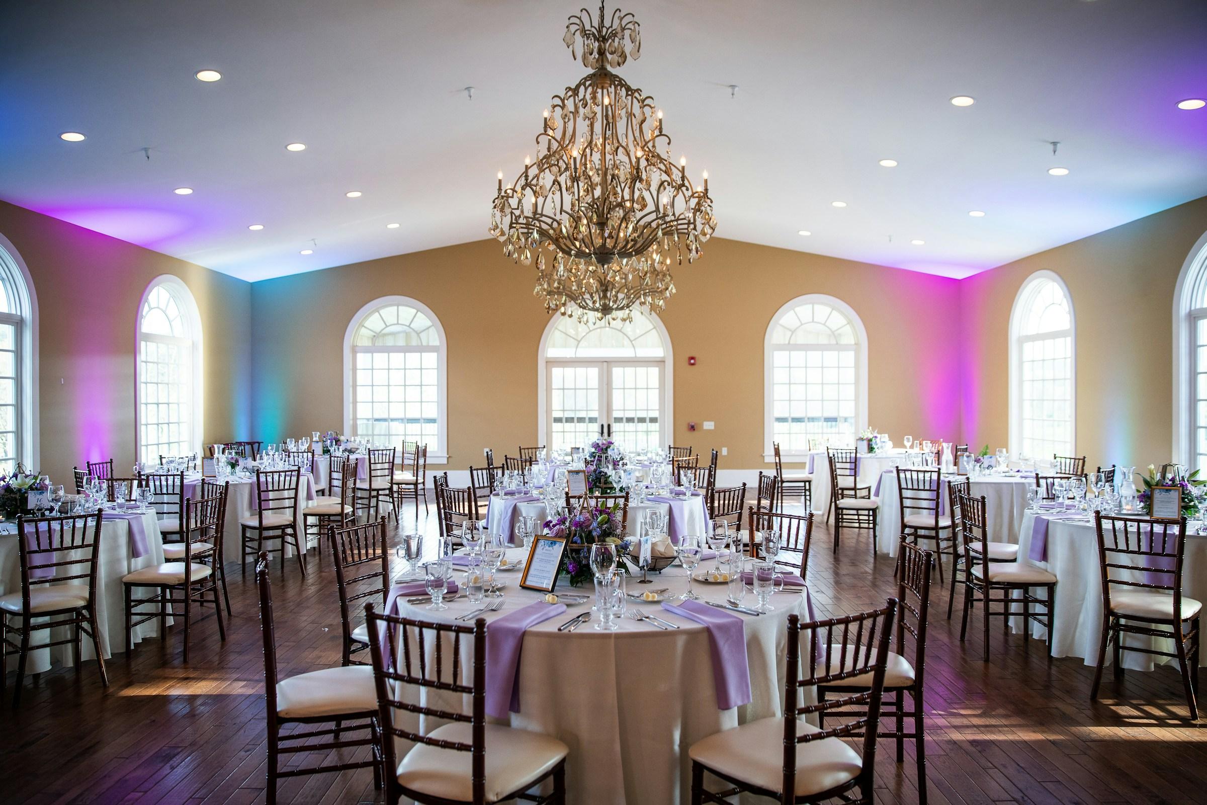 Empty banquet room set for a reception: round tables with white linen, lavender napkins and Chiavari chairs under a crystal chandelier, arched windows and soft pink-and-teal uplighting