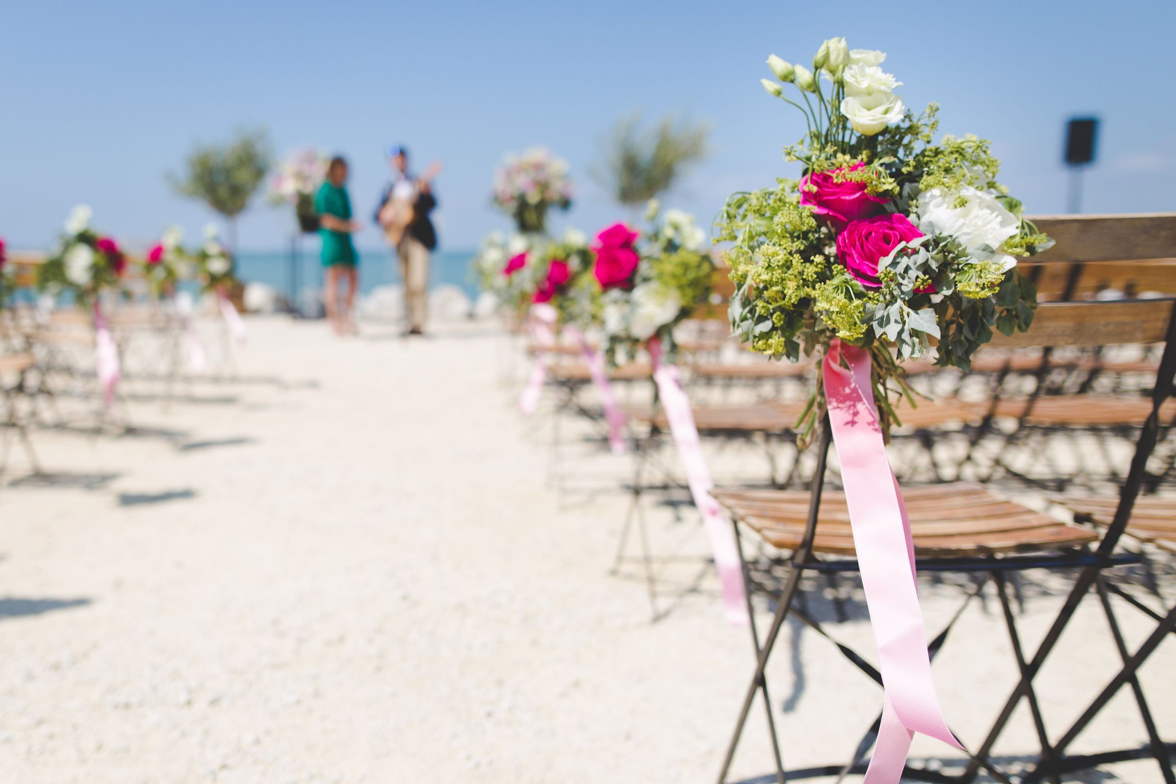 Beach ceremony aisle of wooden bistro chairs, each tied with a pink ribbon and a posy of fuchsia roses, facing the sea under a clear blue sky. Two small, fully blurred figures stand at the far end