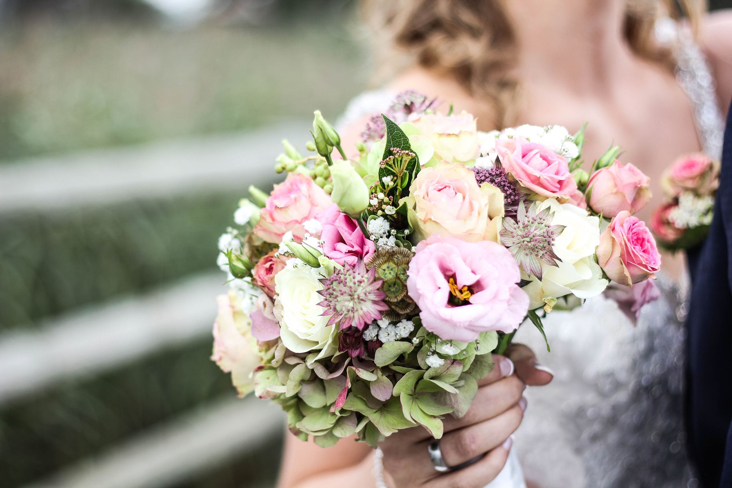 Close-up of a bridal bouquet of pink lisianthus, blush and cream roses, astrantia and green hydrangea held against a lace gown; the face is out of frame and heavily blurred