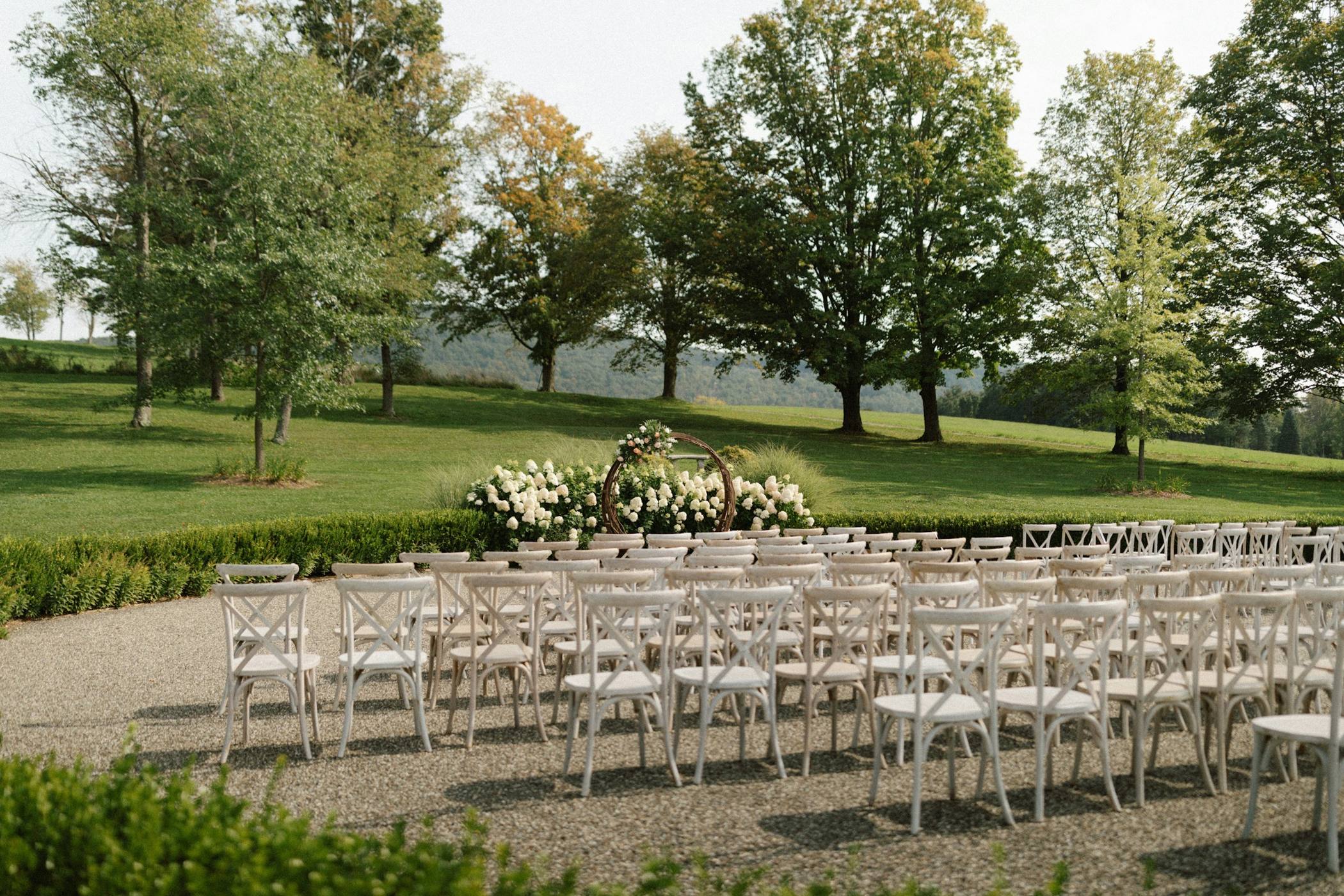 White cross-back chairs lined up on a gravel terrace facing a circular arch of white hydrangeas and roses, with a green lawn and mature trees beyond