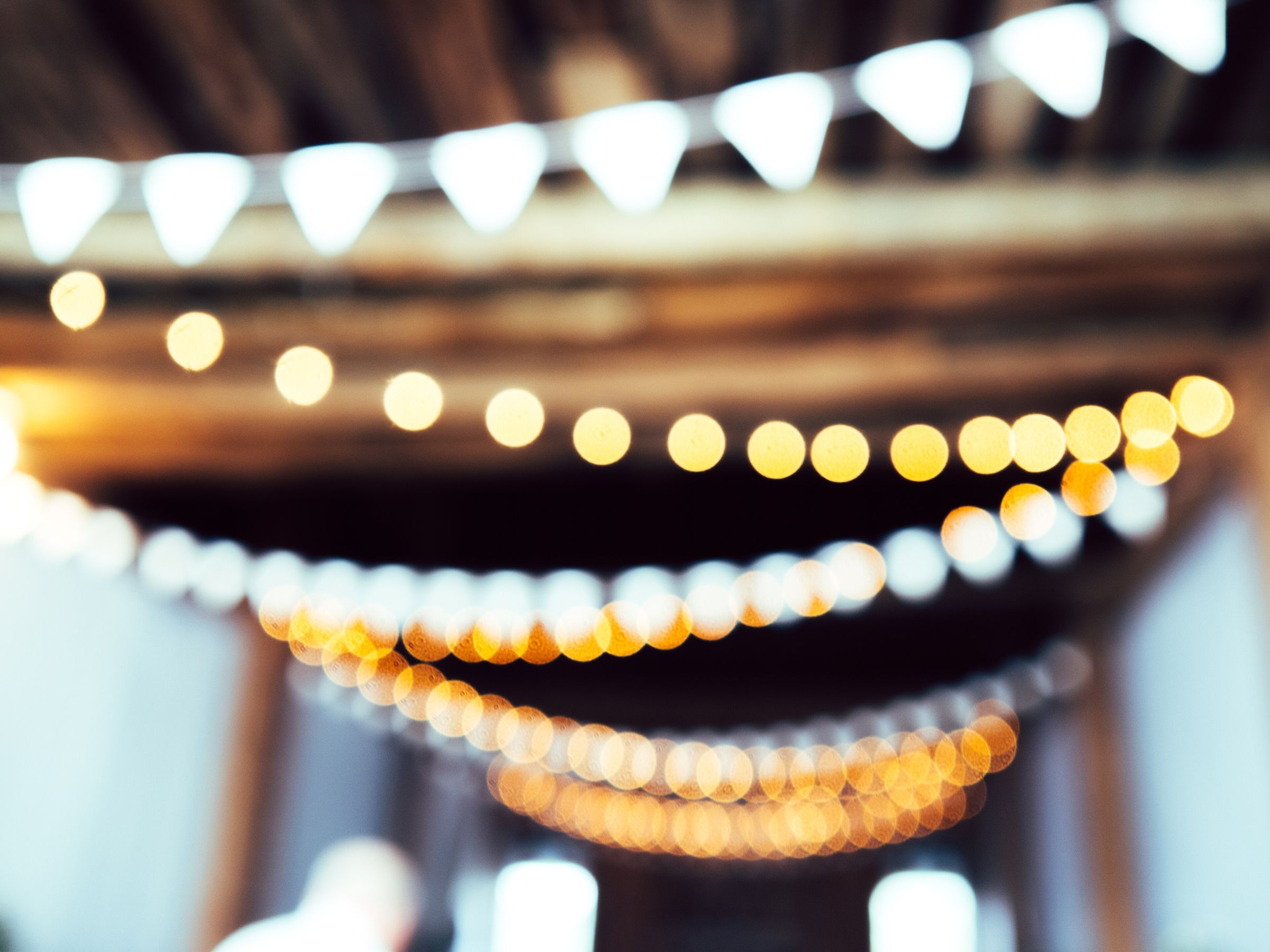 Soft-focus festoon lights and white bunting glowing beneath timber beams: an abstract party-atmosphere shot, usable for dance-floor mood (one blurred shape at bottom left)