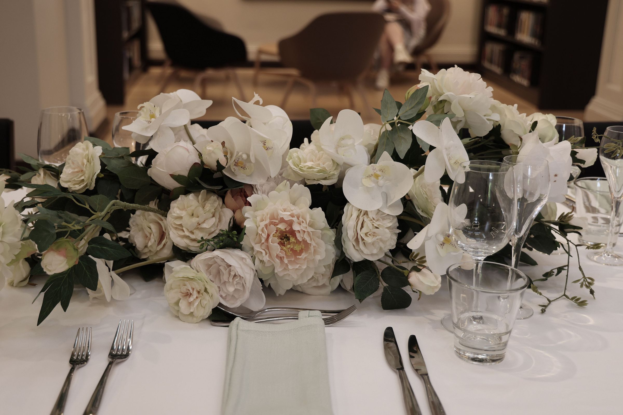 Low centerpiece of white orchids, blush peonies and garden roses spilling across a white-linen table set with silver cutlery and glassware. A blurred seated figure is visible in the far background