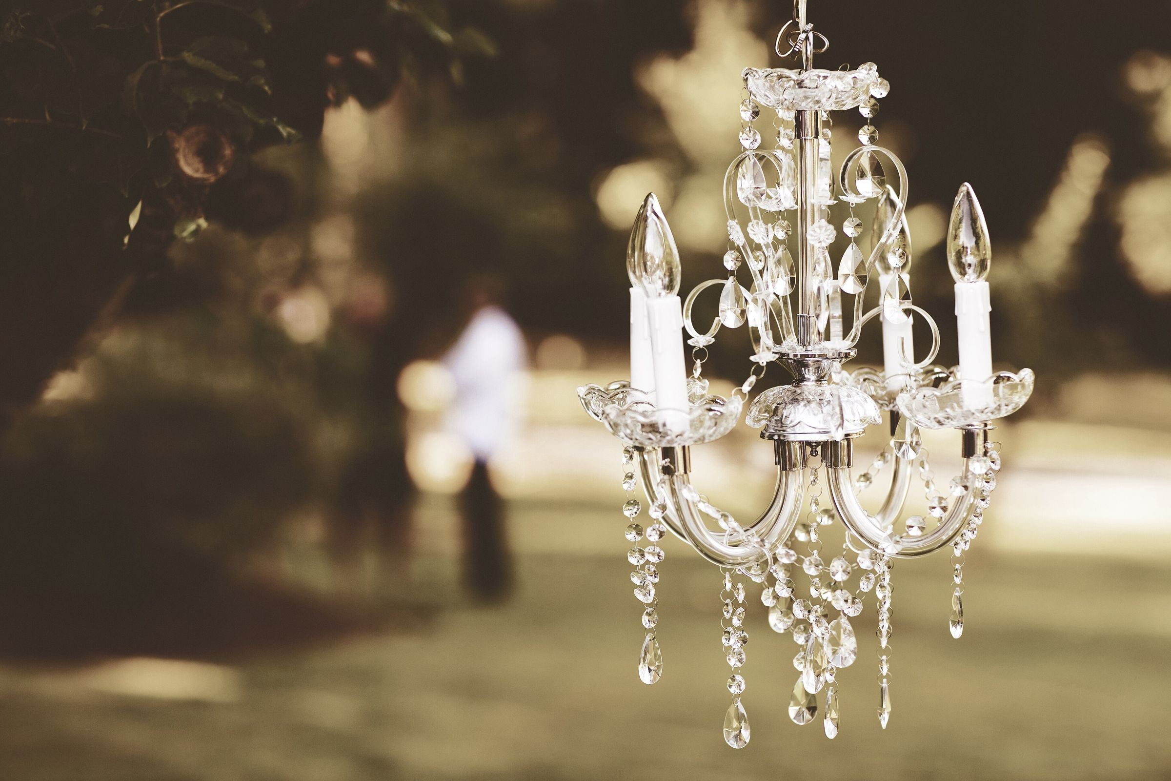 A small crystal chandelier hanging from a tree branch outdoors against a warm, blurred sepia garden (a blurred distant figure in the background)