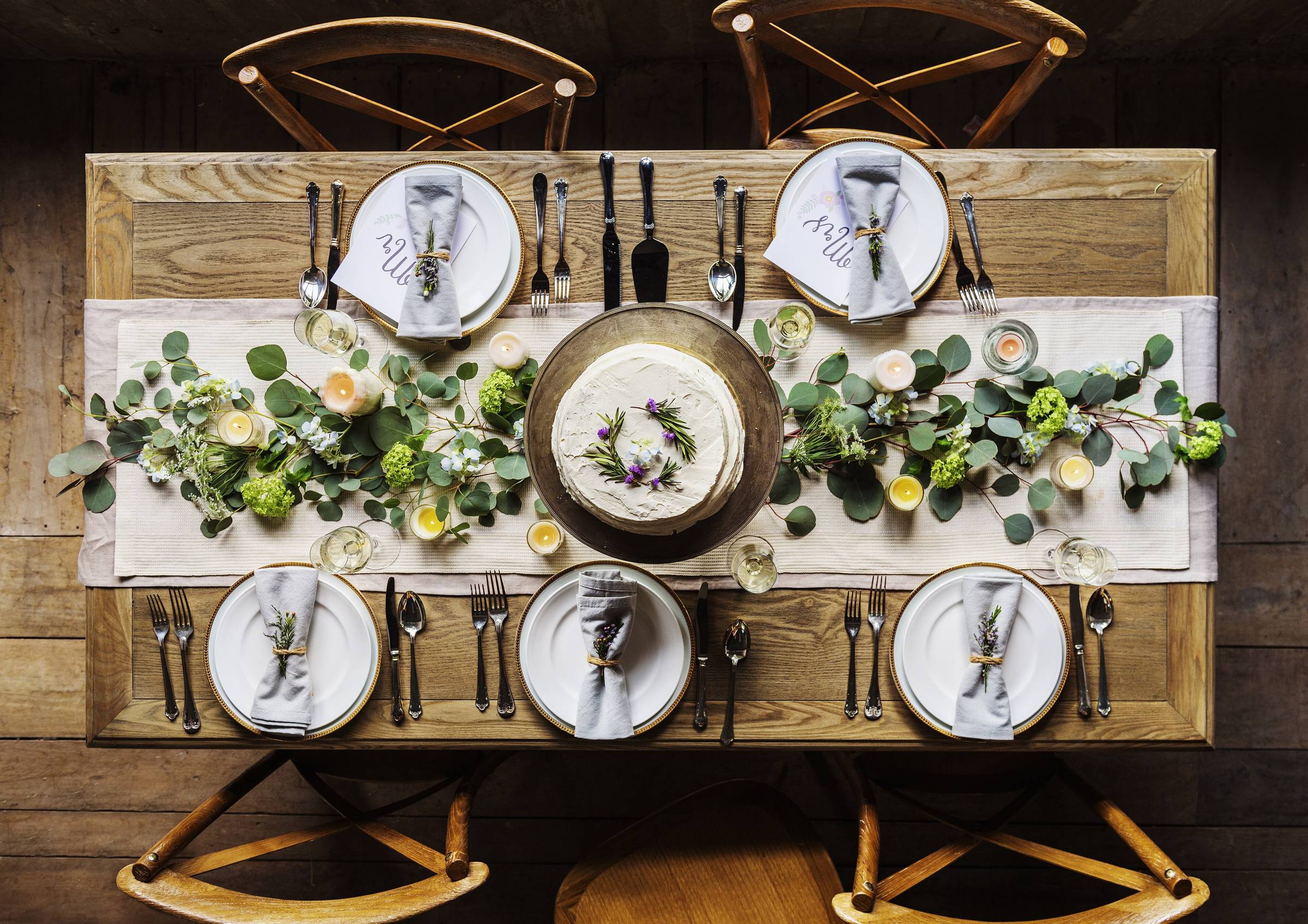 Overhead view of a rustic wooden dinner table with a eucalyptus and hydrangea garland runner, tea lights, a naked white cake at the center and gray napkins tied with herbs at each place