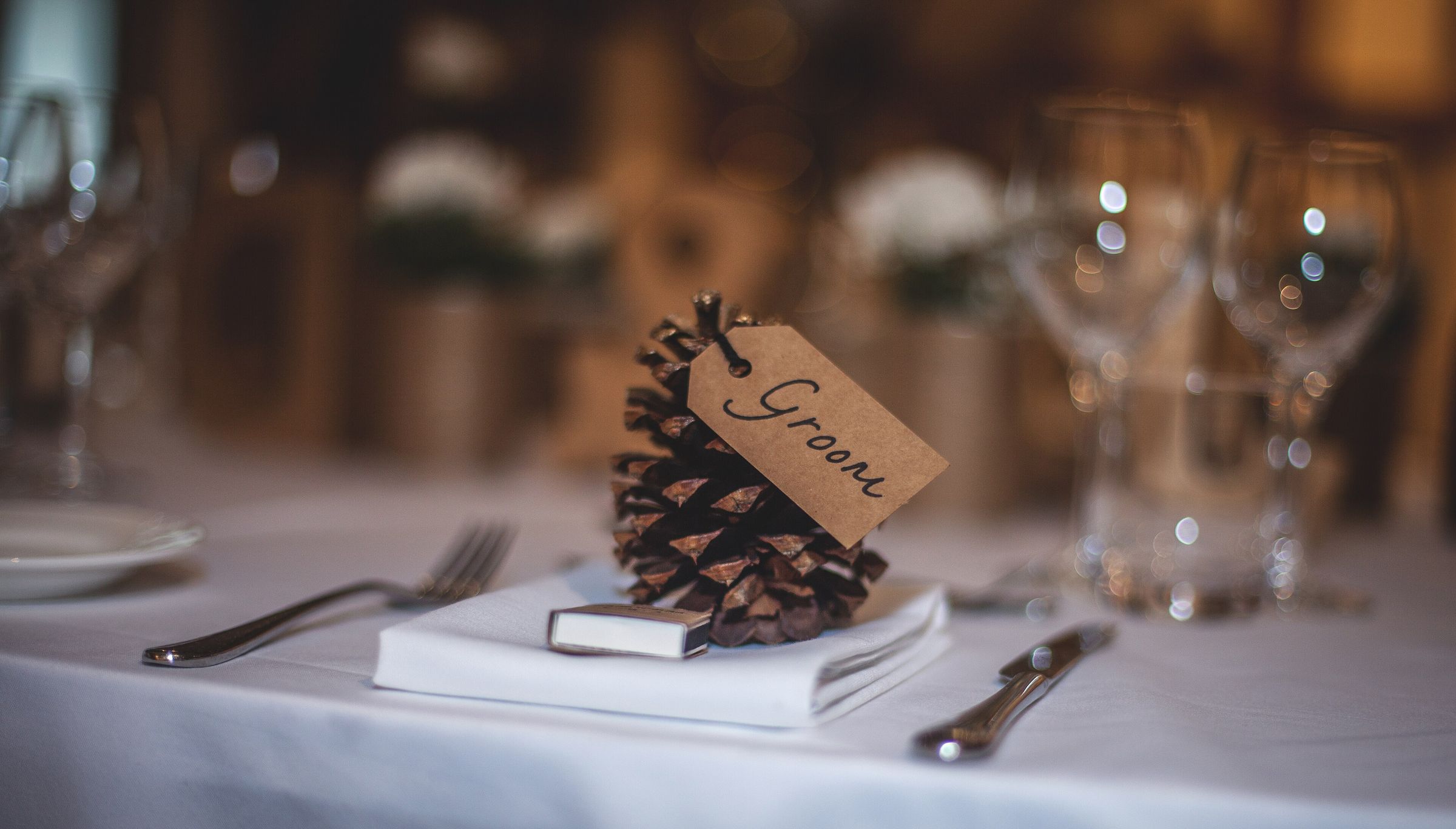 Pine cone place-card holder with a kraft tag hand-lettered "Groom" on a folded white napkin, flanked by silverware, with glassware blurred behind