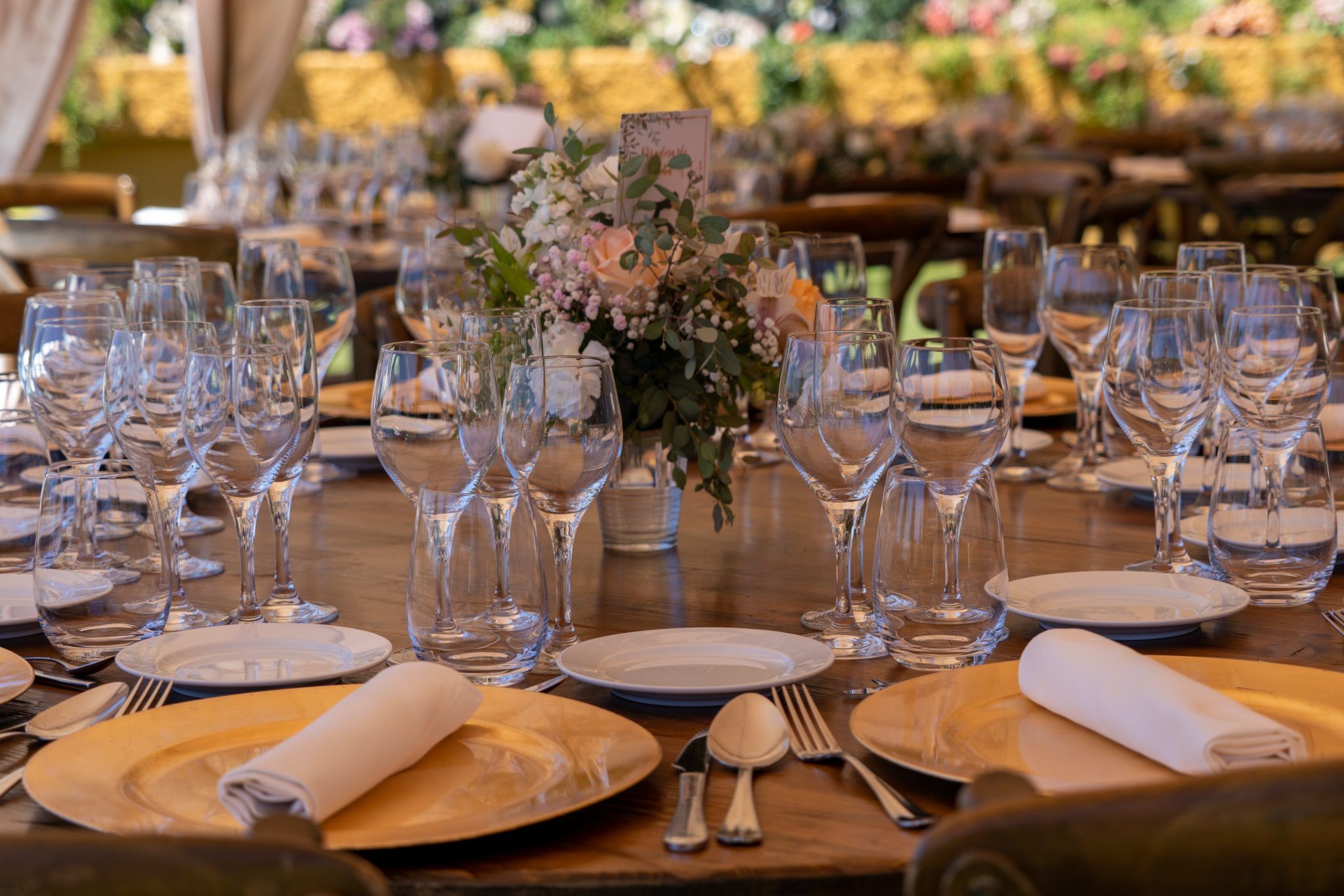 Round wooden reception table set with gold chargers, rolled white napkins, rows of wine glasses and a low arrangement of blush roses and eucalyptus, under a floral-draped marquee