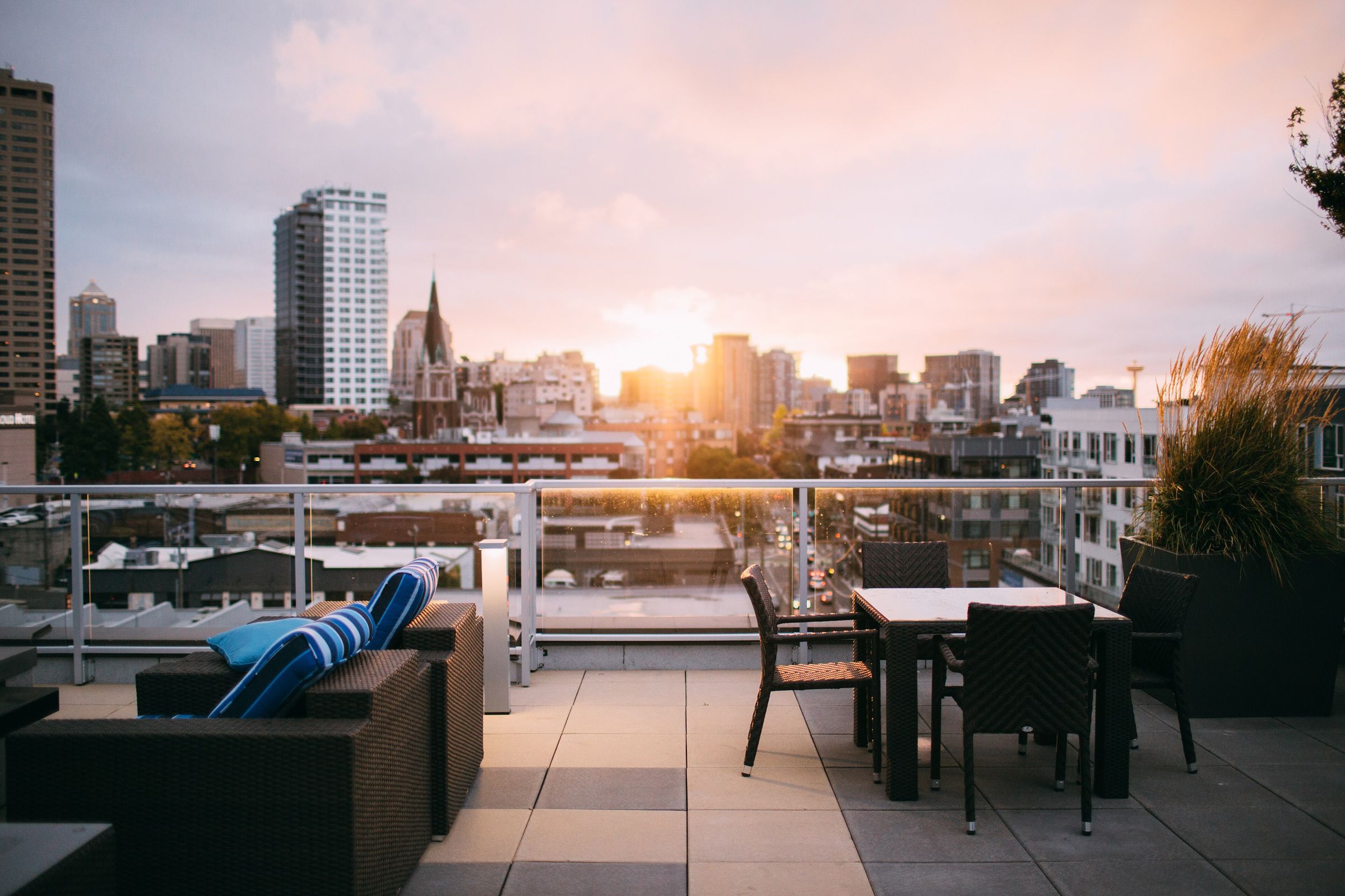 Empty rooftop terrace with wicker lounge seating and a dining set behind a glass balustrade, looking over a downtown skyline at sunset (Seattle; no hotel identifiable)