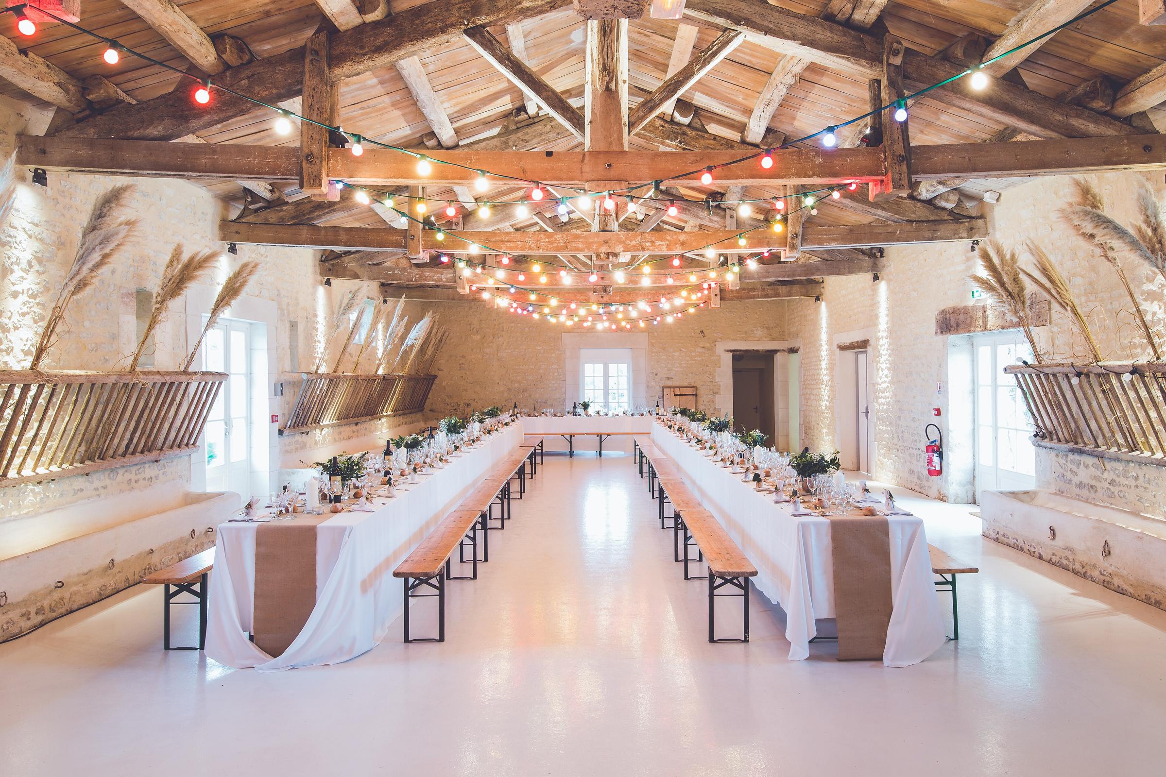 Long reception tables with burlap runners and wooden benches in a whitewashed stone hall, with colorful festoon lights strung across exposed timber rafters and pampas grass on the walls