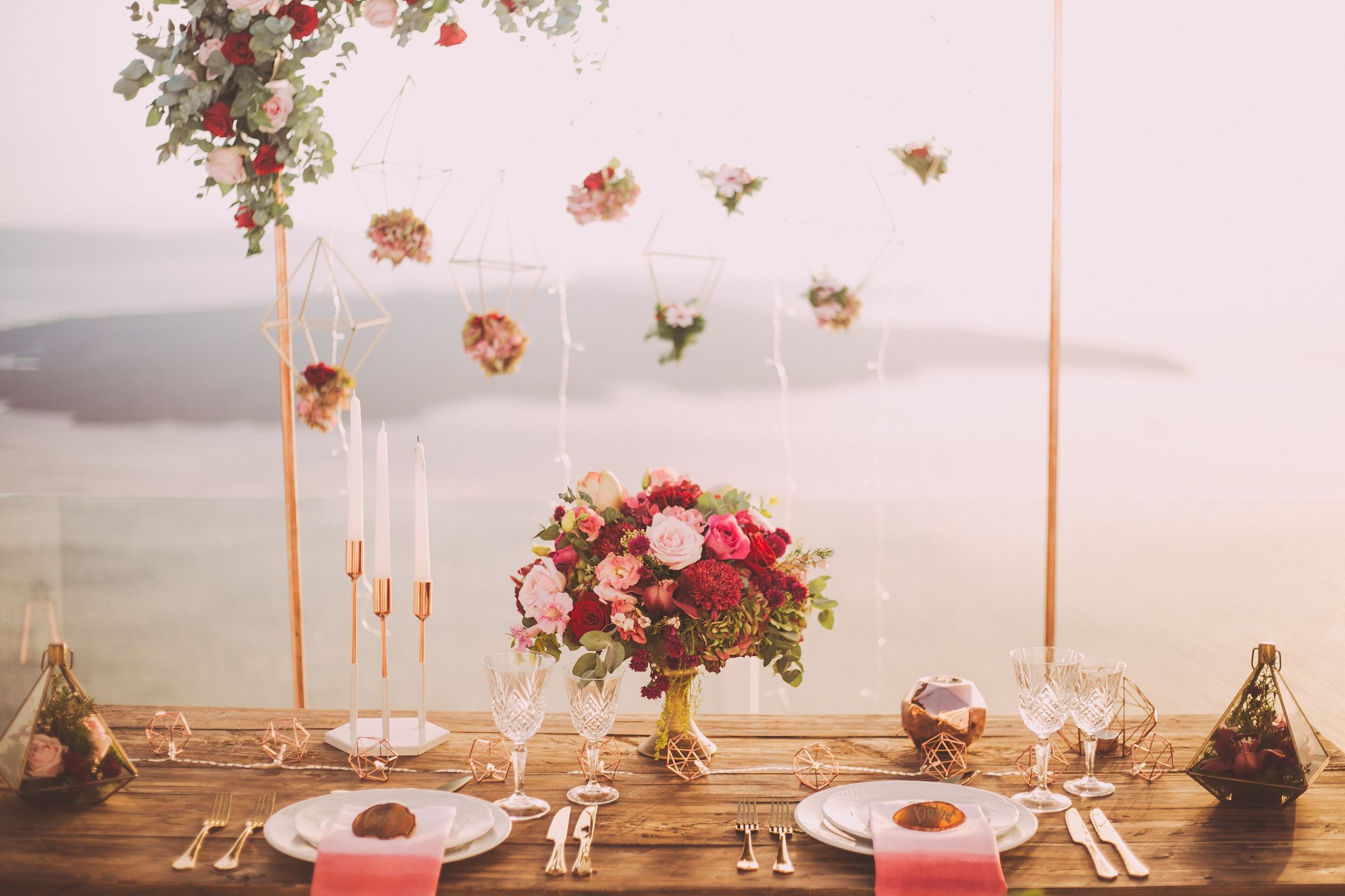 Sweetheart table for two on a clifftop terrace: red and pink rose centerpiece, copper candlesticks, crystal glasses and geometric terrariums, with hanging flowers and a hazy sea and island behind