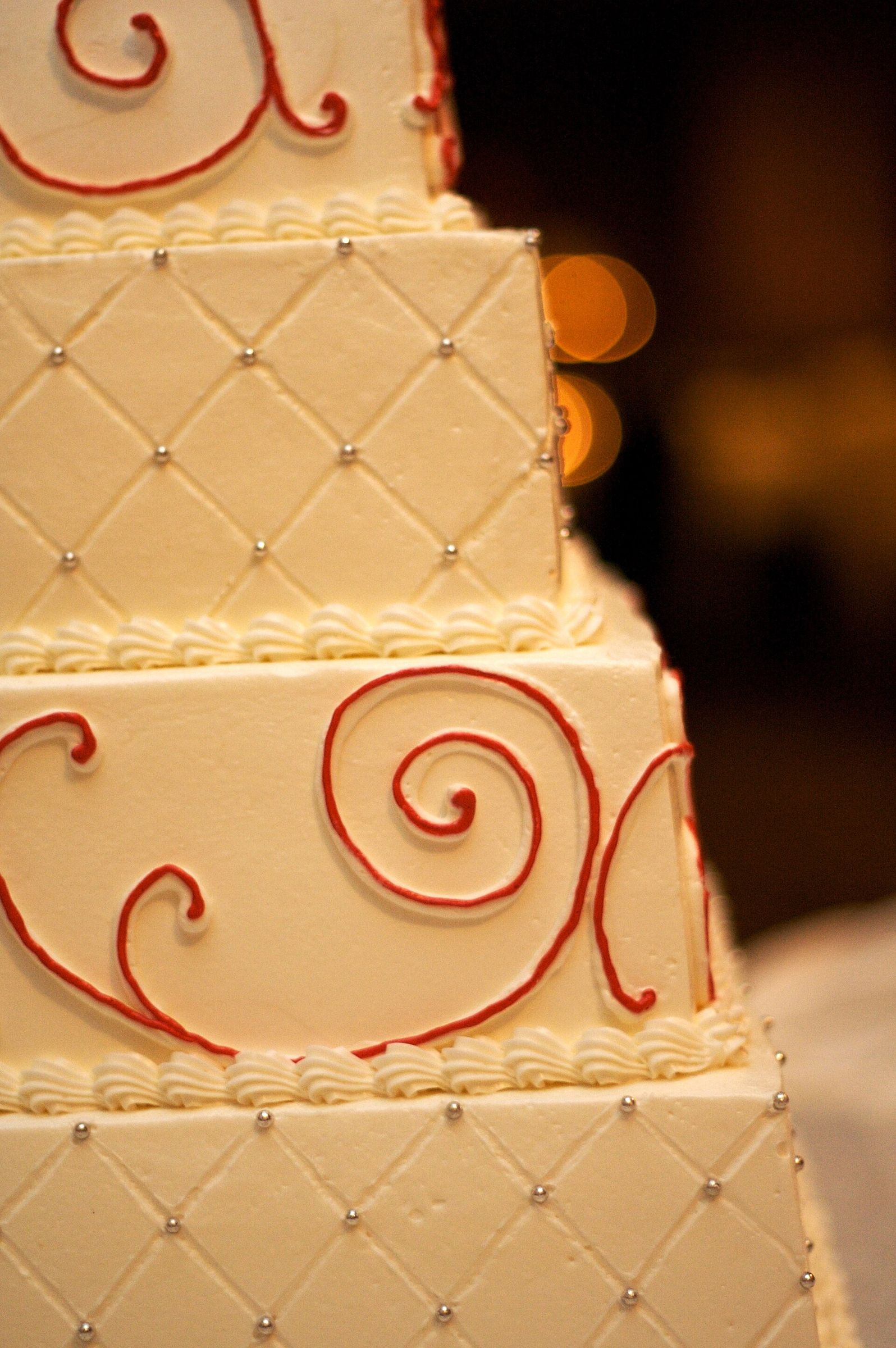 Close detail of a square tiered buttercream wedding cake with quilted piping, silver dragees and red scroll accents, warm bokeh behind (portrait)