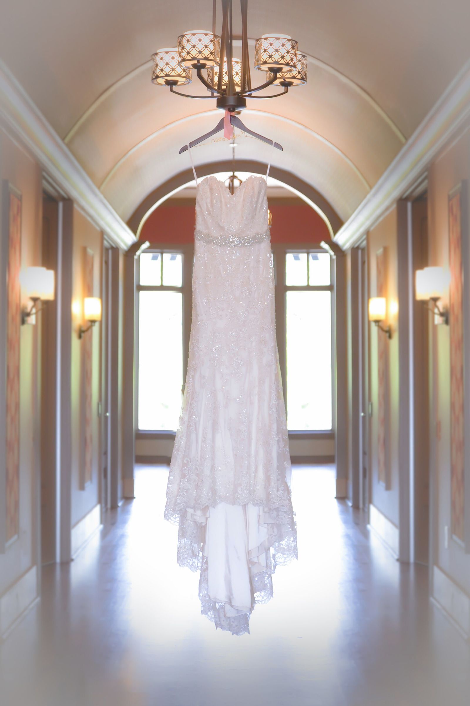 Beaded lace wedding gown hanging from a chandelier in a softly lit barrel-vaulted corridor with wall sconces and tall windows (portrait, no people)