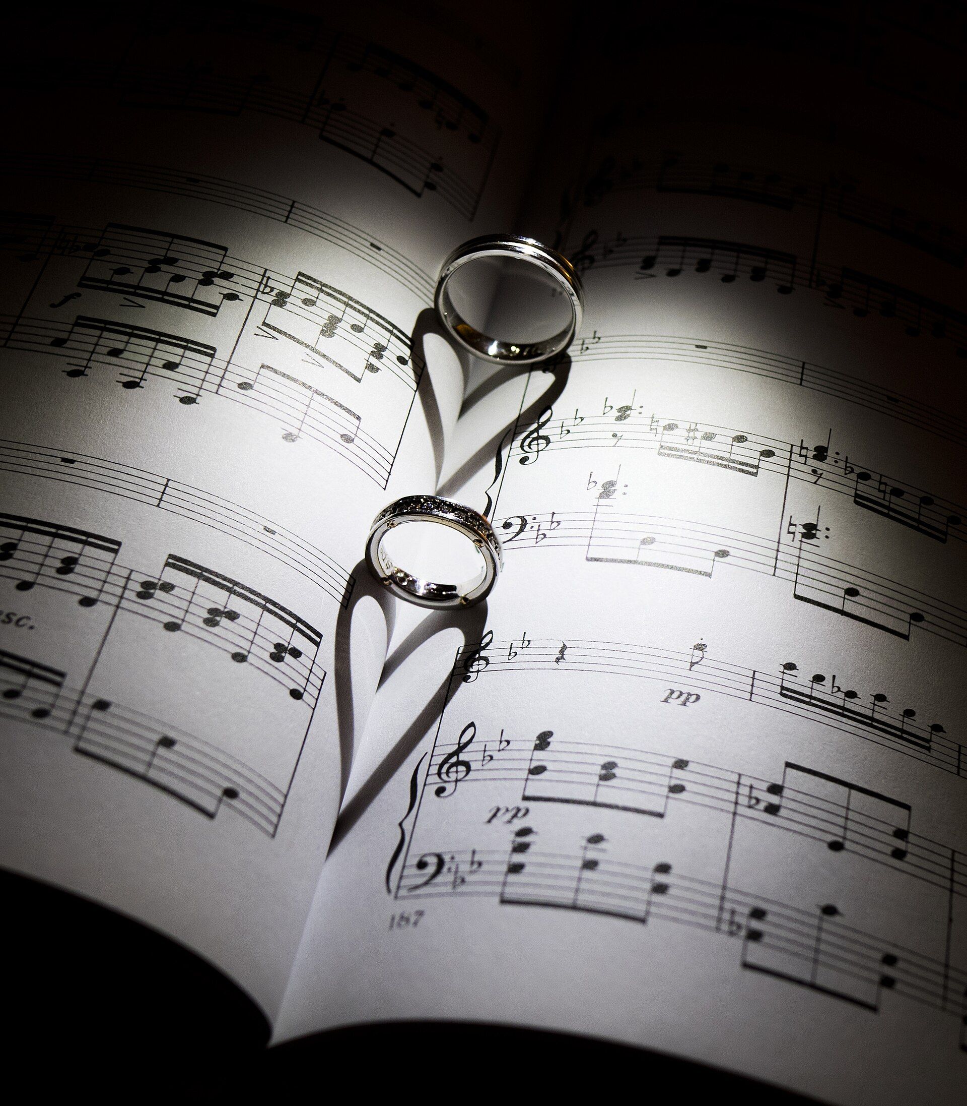 Two wedding bands standing in the fold of an open music score, casting heart-shaped shadows under a single spotlight (portrait)