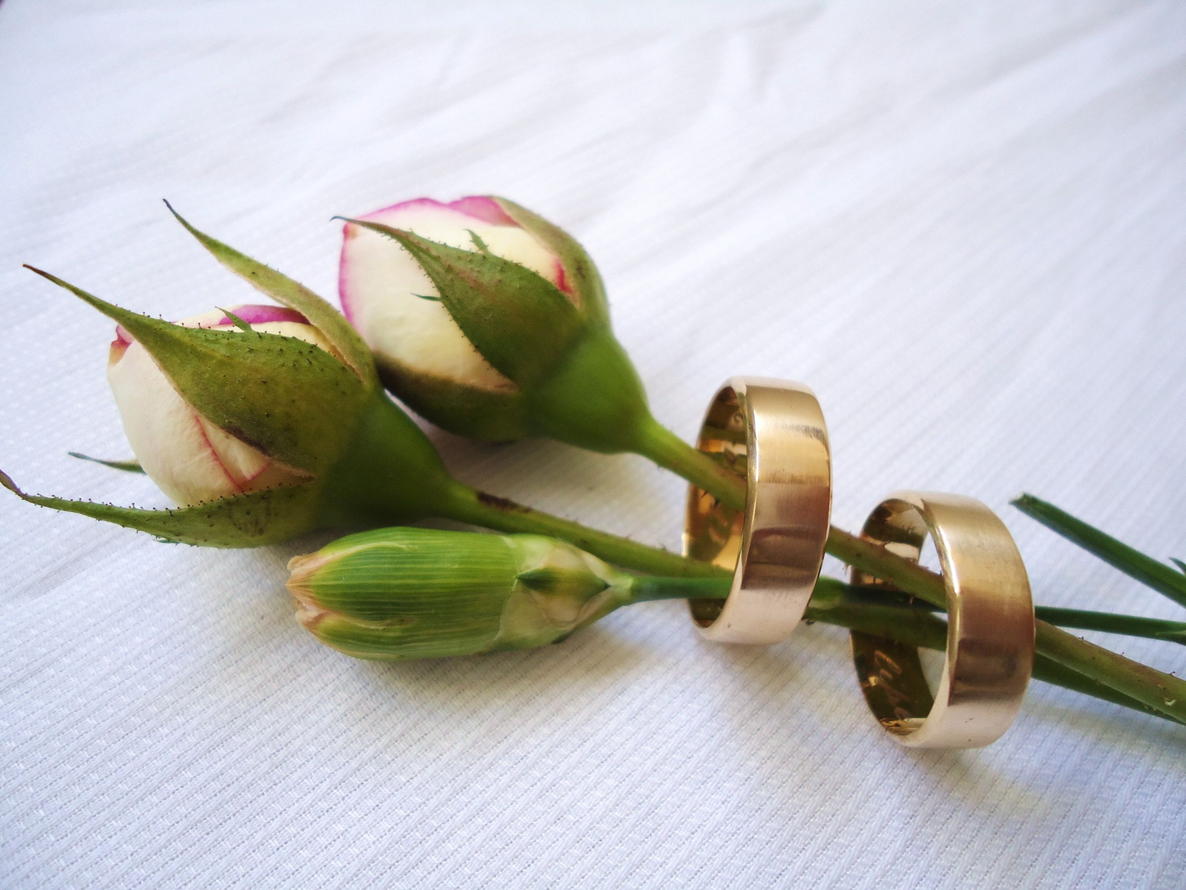 Two gold wedding bands threaded onto the stems of pale pink rosebuds and a carnation bud on white textured linen